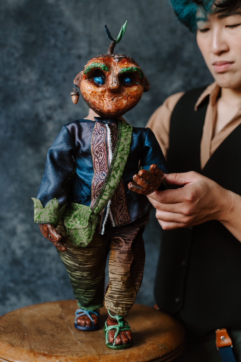 A Taiwanese female-presenting artist with black hair, dark eyes poses with a small humanoid puppet. The puppet has brown skin and a tree branch emerging from its head.