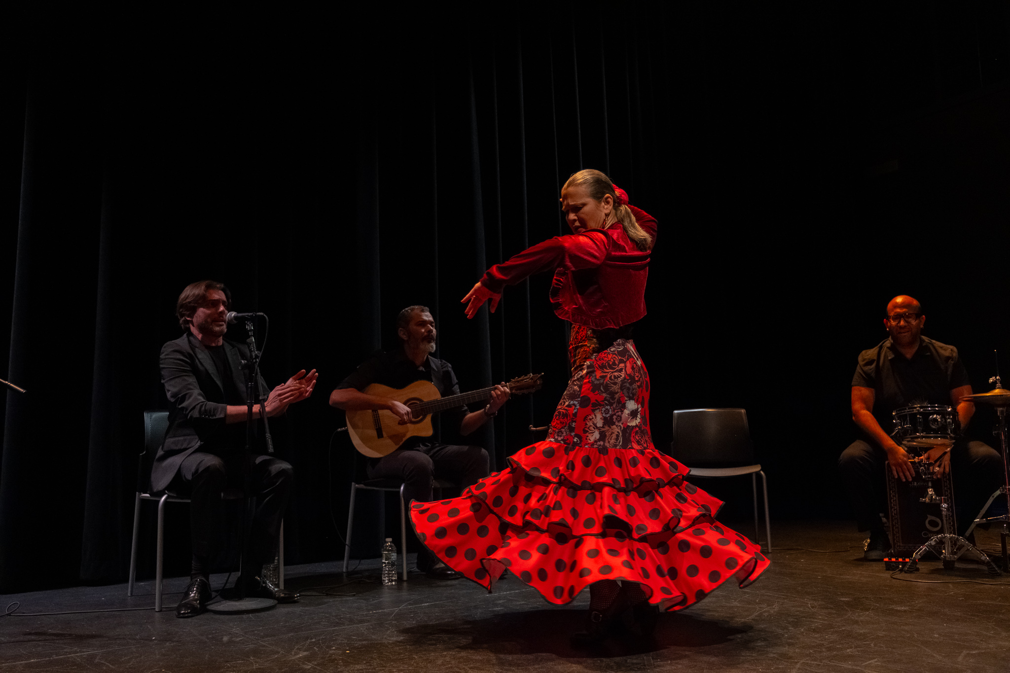 A white woman with grey/brown pulled back hair dances flamenco on stage. She is wearing a long red dress with black polkadots on the skirt and a red flower in her hair.