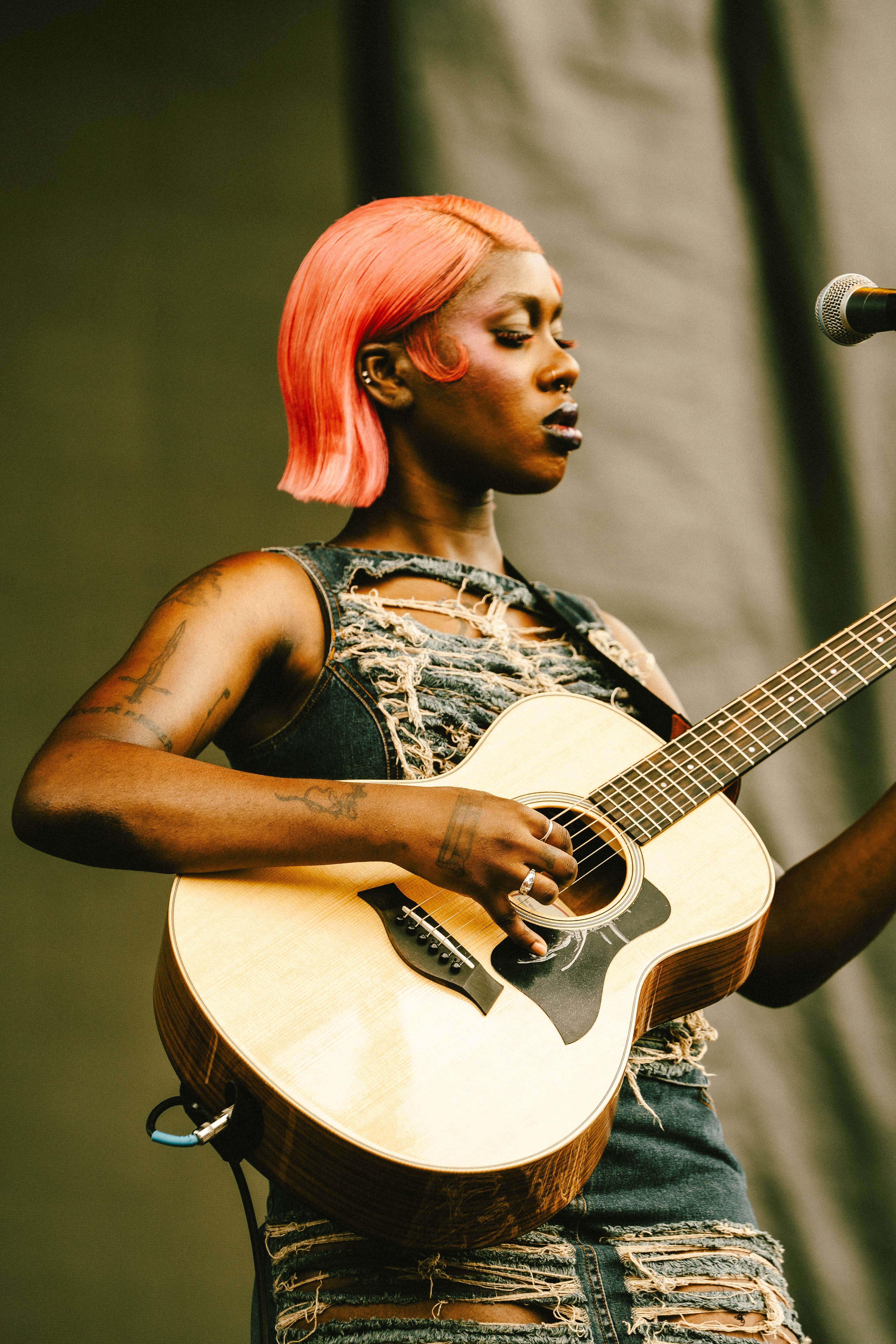 A Black girl with tattoos playing the guitar. She has short orange hair and is wearing a torn denim dress.