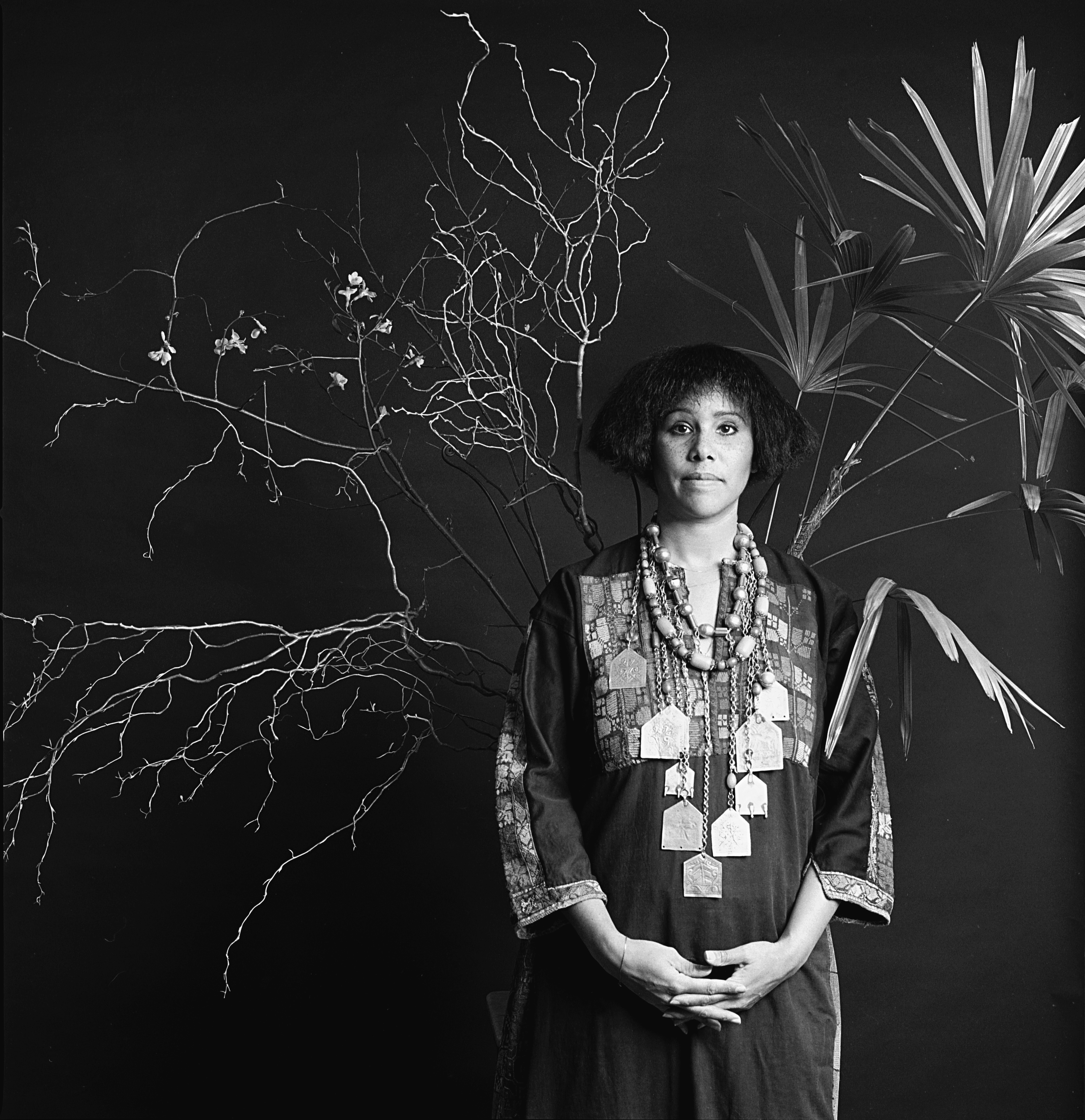 A black and white portrait of the artist. They are wearing a chunky necklace with a patterned dress. Behind them are twigs.