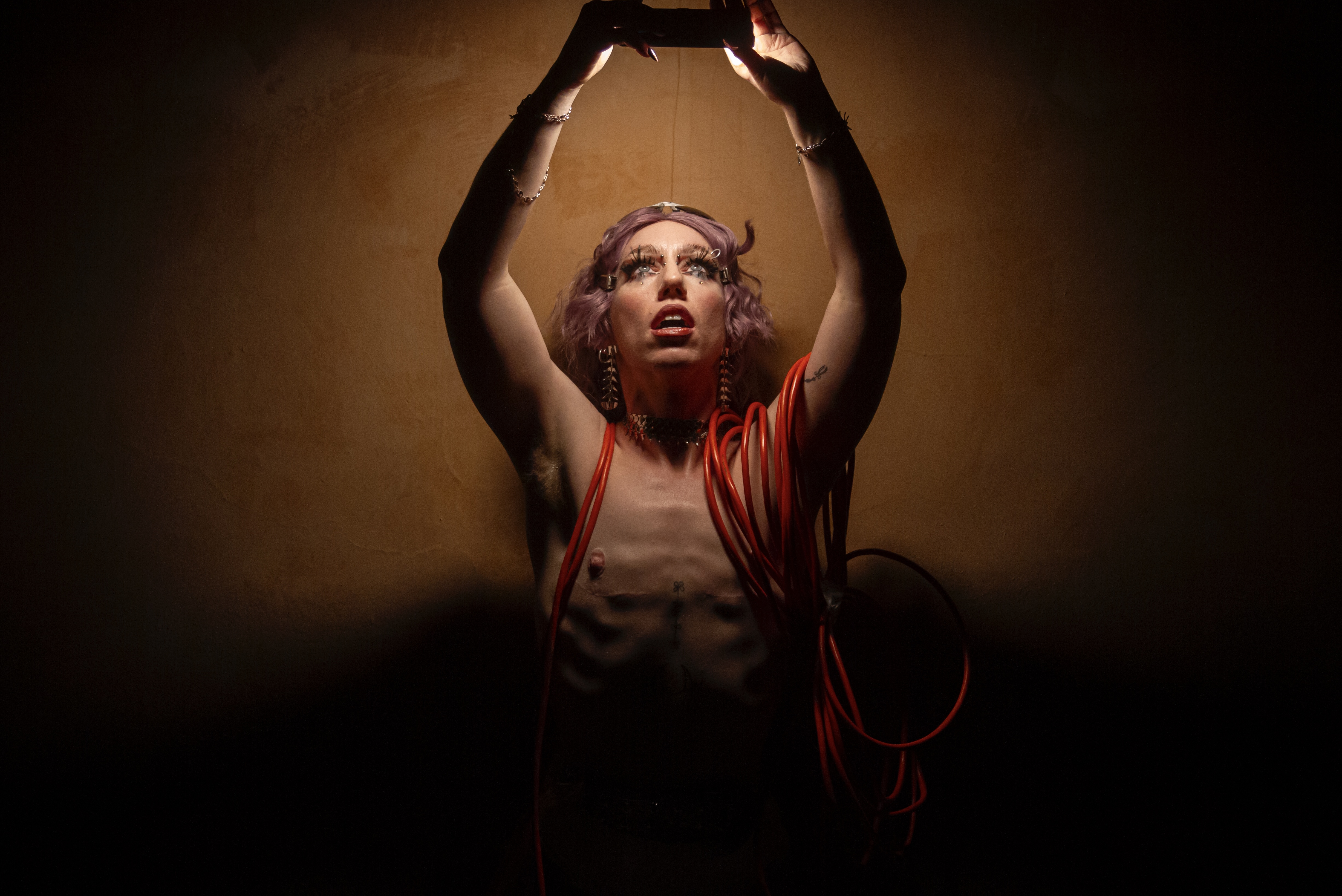 A queer non-binary performer stands against a wooden wall. They are lit by the light of a phone that they are holding above their head. They have purple hair and are shirtless, wearing red electrical wire and dangling jewelry.