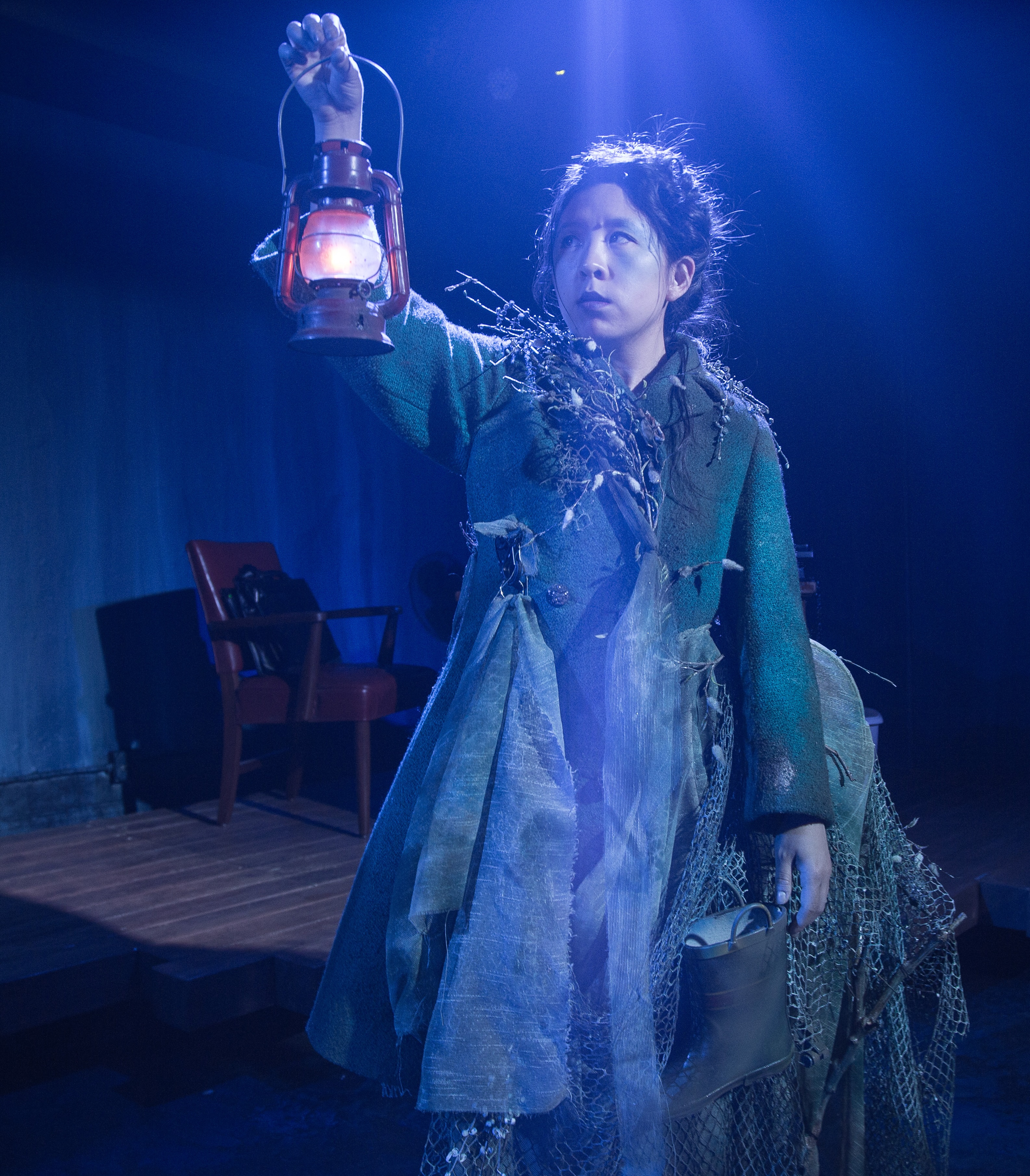 A performer stands on stage holding a prop lantern. They are wearing a green and blue outfit and has their hair tied up in a bun.