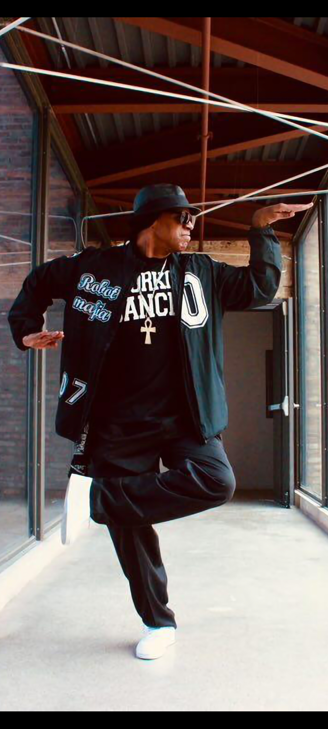 A Black man with sunglasses and hat poses with his hands extended in the middle of dancing.