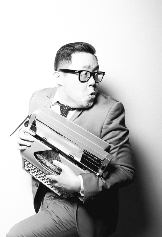 A black and white image of a Filipino-Chinese cis male with short dark hair, black glasses and olive skin poses with a typewriter.