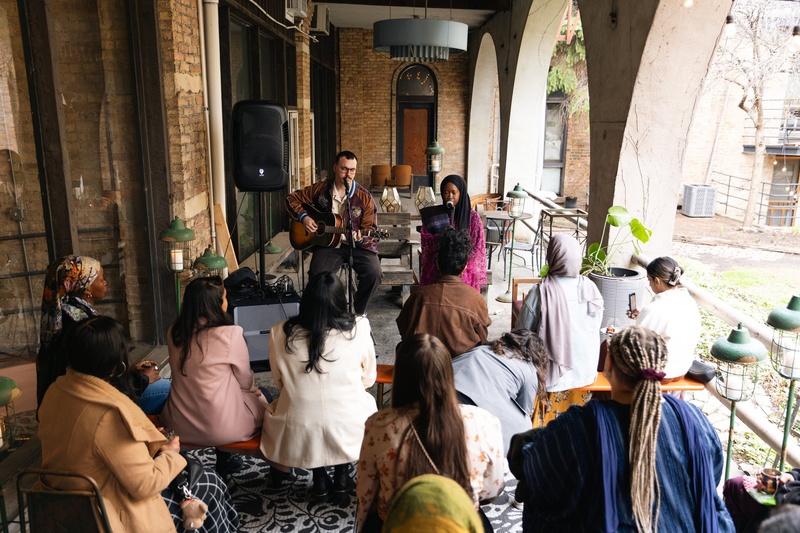  A Black Woman in a Hijab sits at a microphone. She is wearing sa pink dress. A person is next to her sitting on a stool playing the guitar.  You can see members of the audience from behind sitting in front of them. 