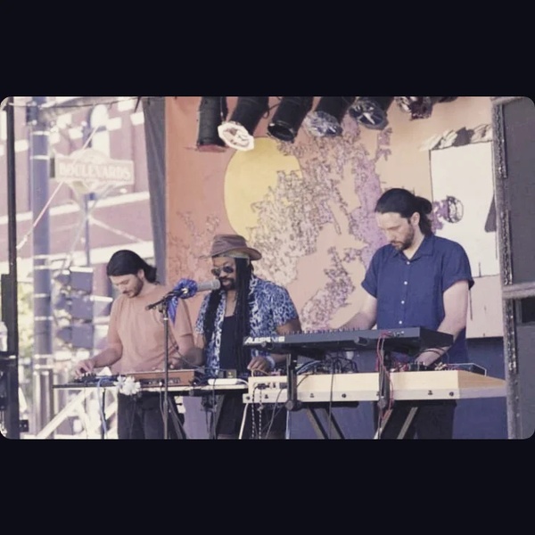  A Black/queer performer with Dreadlocks stands at a microphone, singing. There are two other performers flanking them. They are outside on a festival stage. 