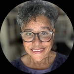 A headshot of a black woman with freckles, dark rimmed glasses and grey culy hair.