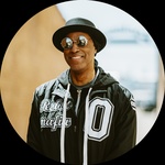 A Black man with sunglasses and hat smiles at the camera.