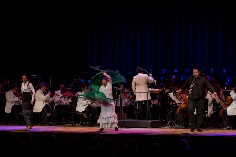  A white woman with grey/brown pulled back hair dances on stage while manipulating a green fringed piece of fabric. She is wearing a white dress and is surrounded by an orchestra. 