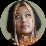 Filipinx femme person with layered black hair with blonde in the front poses with flowers in front of a green background.