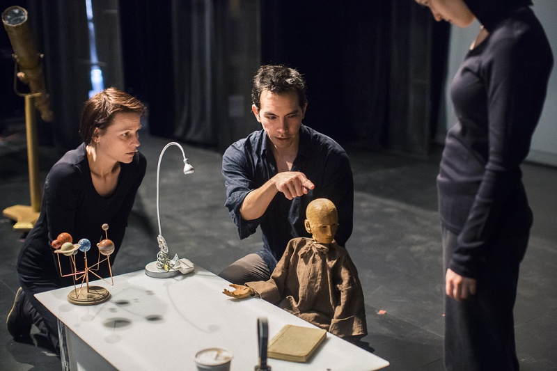  a biracial person with long black hair shows others how to operate a basic human puppet made of wood. 