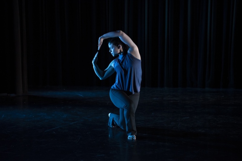  A white woman with red hair tied back performs on a dark stage. They are holding their arms in a circle motion to the side of her body. 