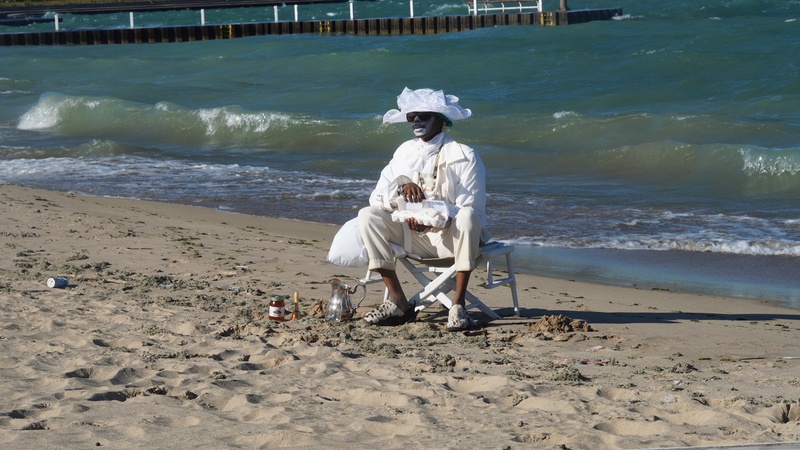  Performer sits in the sand on a beach. They are wearing an all white outfit and holding a box of eggs. 