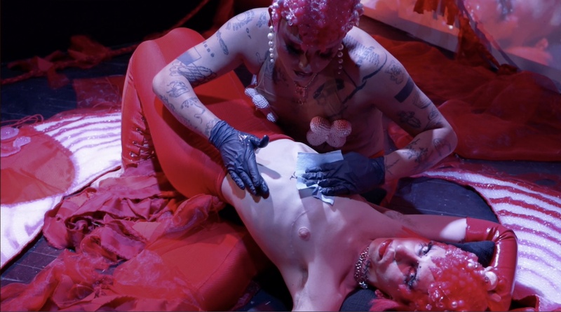  Still from a performance where a non binary performer is getting tattooed on their stomach. They are wearing a red outside and looking straight into the camera while the other performer tattoos their stomach. 