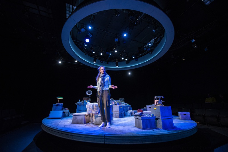  A performer stands on a circular set filled with boxes. They are wearing a white shirt and brown pants. 