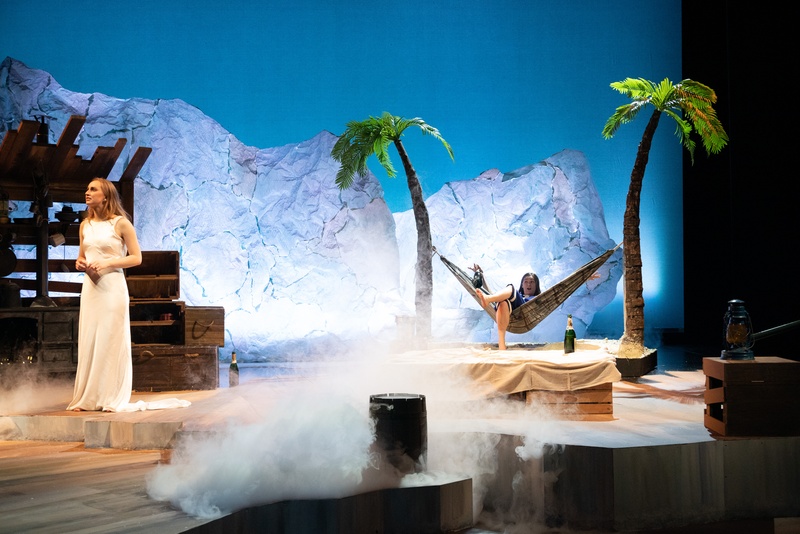  A performer stands on a set that looks like it is on the beach. One performer is in a hammock. 