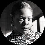 A black and white portrait of the artist LOLA AYISHA OGBARA. She is standing in a brightly lit studio. She has black skin and slicked back hair.