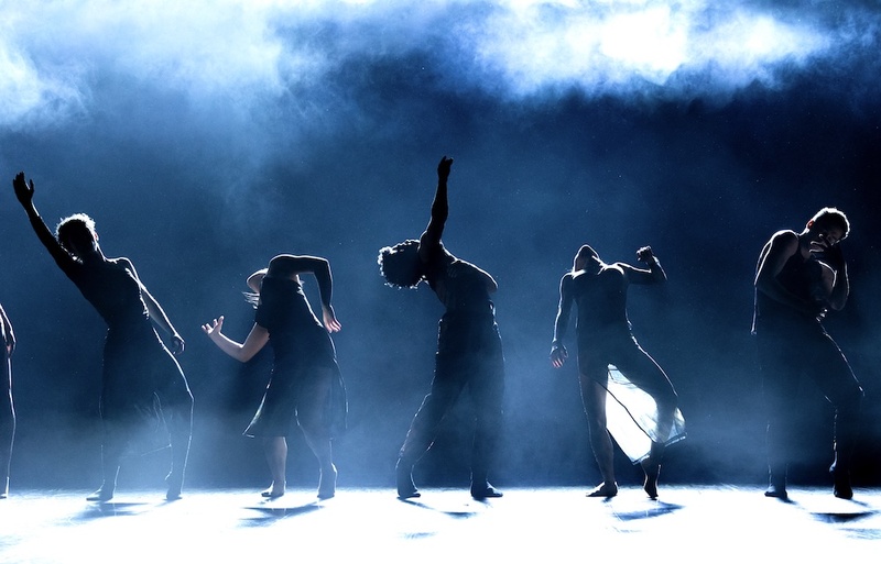  Five performers dance in an all black room with hazy blue fog in the air. The figures are obscured by the lighting in the room, casting shadows on all of them. 