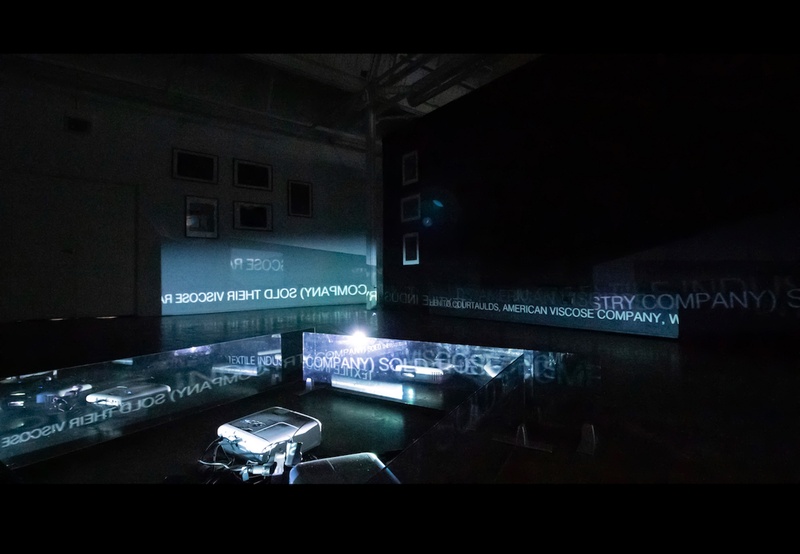  A dark room lit by two projector on the floor. The projectors are displaying words on to two way mirror panels. 