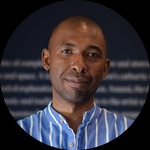 Black masc presenting person with a bald head in a blue shirt standing in front of a painting.