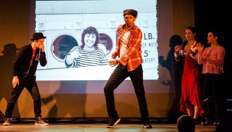  Four people perform on a stage with a projected photograph behind them. One individual is wearing flannel and in dancing in front while two people are clapping to the side. 