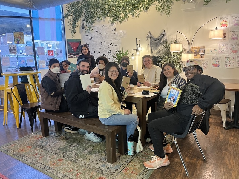  A large group sits around a table. They are all smiling towards the camera and each person is holding a small chapbook. They are in a room with a big window and white walls. 