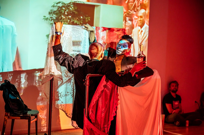  Two performers face each other while holding up a sheer fabric that is being projected on. One performer holds a bottle of liquor while the other has white face paint on. 