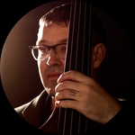 A white man with glasses looks off to the left as he holds on to a bass.