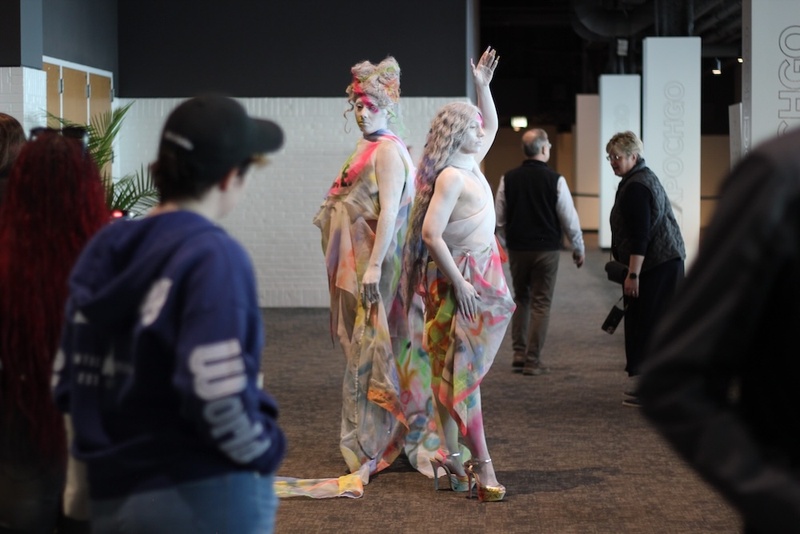  Two performers covered in white paint stand at Art EXPO. They are posed back to back and wearing multi-colored wrap like garments. 