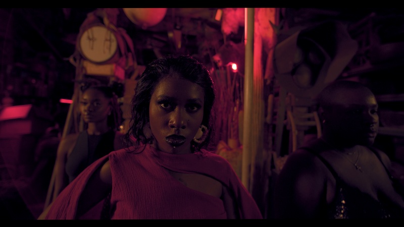  A Black girl with tattoos stares into the camera in a still from a music video. She is wearing large hoop earrings and a red turtlenecked shirt. 