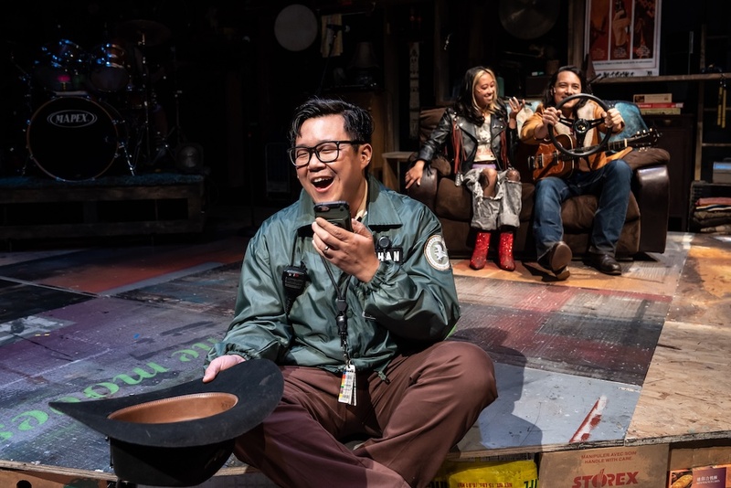  A  Filipino-Chinese cis male with short dark hair and olive skin sits on stage. He is wearing a green bomber jacket and brown pants. he holds a black cowboy hat and is speaking into a phone. 