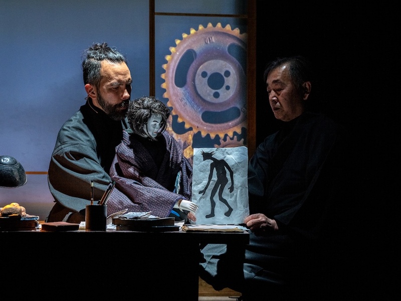  a biracial person with long black hair operates a humanoid puppet of a woman who is standing behind a table. 