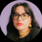 A headshot of a Woman with long brown hair, glasses and facial piercings on her in front of a purple background.