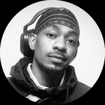 A headshot of a black/queer person in black and white. They have a wrap on their head and over ear headphones dangling from their head.