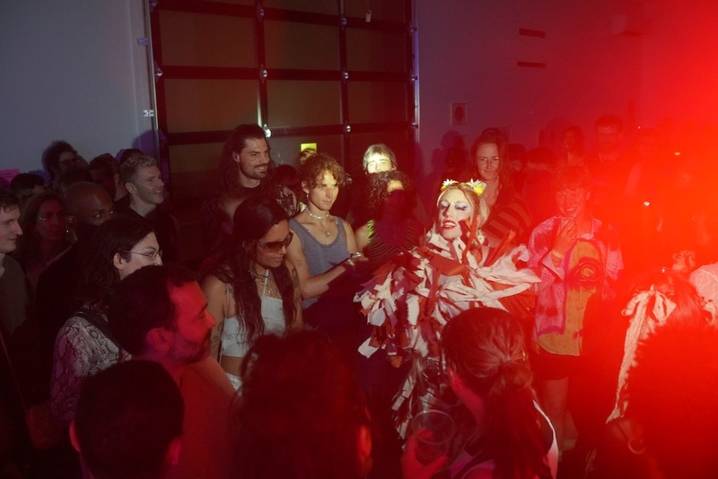  A still from a performance. A non-binary performer stands in a crowd posting during a performance. 