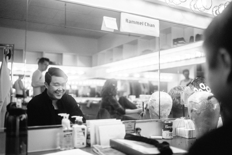  A Filipino-Chinese cis male with short dark hair and olive skin smiles in the mirror in a black and white photograph backstage. 