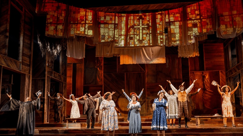  A still from a theater production of The Color Purple. Ten performers hold their hands up in a set made to look like a chuch with projected stain glass windows. 