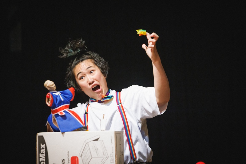  A Taiwanese female-presenting artist with black hair, dark eyes performers with a small humanoid puppet. They are wearing a white button down shirt and rainbow suspenders. 