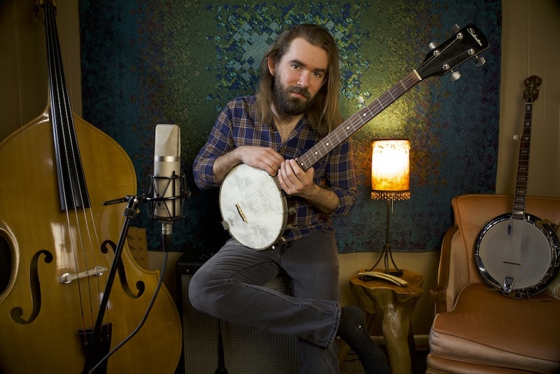  A medium built white man with long, light-brown hair and a dark brown, medium length, full beard poses with a banjo. 