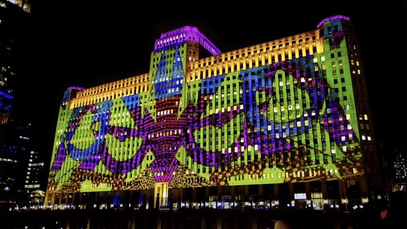  A large building lit up at night. On the building there is a projection of shapes in purple, blue, red and yellow. 