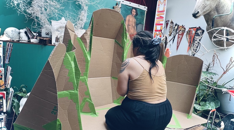  A Filipinx femme person with layered black hair with blonde in the front sits in a cardboard installation. 