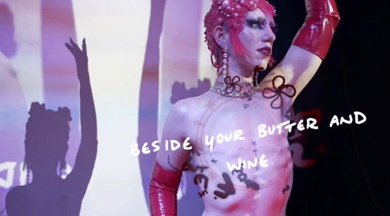  Still from a performance. A non-binary performer stands in front of a wall that is being projected on. They are wearing a red swim cap, red gloves and the words Beside your butter and wine are projected on them. 