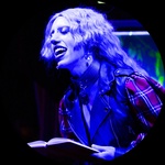 A queer person with curly hair cut at an angle smiles and looks to the side. They are wearing a plaid jacket and are lit in blue.