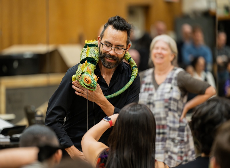  a biracial person with long black hair and glasses operates an iguana puppet that is perched on his shoulders. 