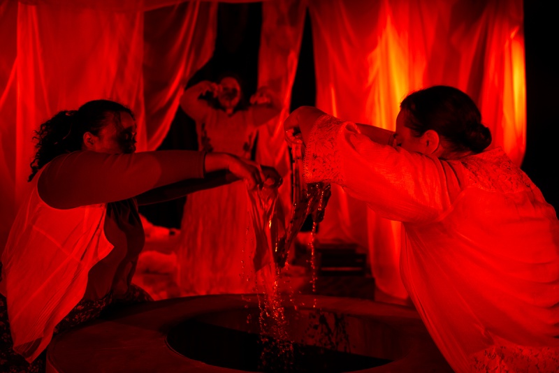  Three performers pull pieces of fabric from a well. The water is dripping off the fabric. The entire scene is saturated in red light. 