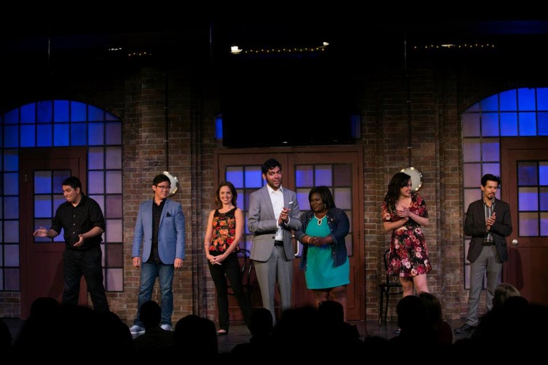  Seven people stand on a stage. Behind them are blue lit windows. Some people are clapping. 