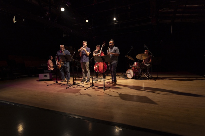  A six piece band stands and plays in a dark room with light wooden floors. 