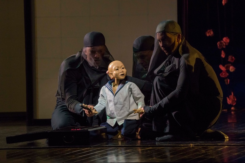  Three puppeteers dressed in all black with black over their faces operate a human like puppet that is wearing a blue sailor shirt. 