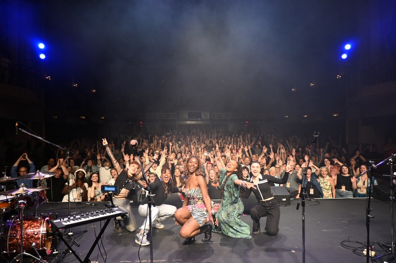  A Black girl with tattoos posses with her band on stage with the entire audience. 