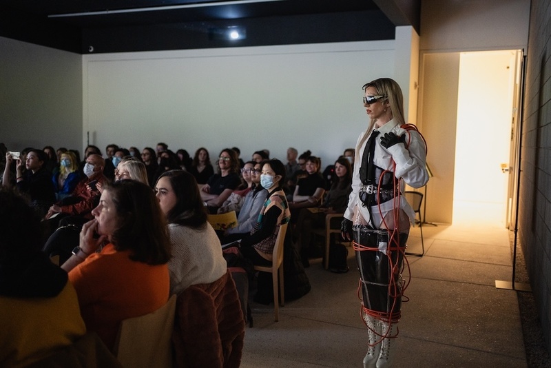  A non-binary performer is standing in an audience. They are wearing a white button down school girl like outfit and are wrapped in red electrical chord. 